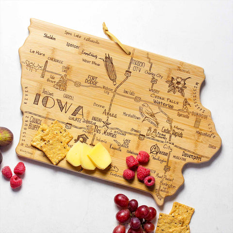 The Destination Iowa Cutting Board by Totally Bamboo, measuring 15-1/2" x 10-1/4", features a bamboo surface shaped like Iowa with laser-engraved city names and landmarks. It is elegantly arranged with cheese, crackers, raspberries, and grapes on the side.