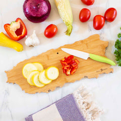 A variety of fresh vegetables, including a red onion, cherry tomatoes, corn, red pepper, garlic, and sliced yellow squash, are arranged on a white marble surface. Nearby is the Totally Bamboo Destination Puerto Rico Cutting Board (18" x 7") with a knife and chopped red pepper resting on it alongside a folded towel.