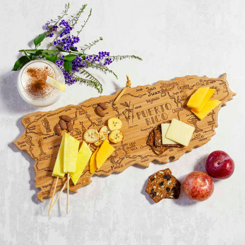 The Totally Bamboo Destination Puerto Rico Cutting Board, measuring 18" x 7", is beautifully designed as a wooden charcuterie board shaped like Puerto Rico from the Destination Series. It features an engraved map adorned with assorted cheese, crackers, and fruit. Accompanying the board is a small dish filled with cinnamon and sprigs of purple flowers to enhance the presentation. Cheese cubes on skewers are complemented by two round fruits.