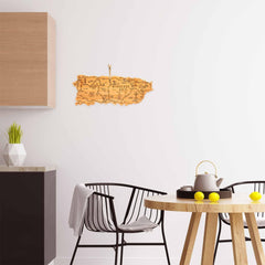 A modern kitchen and dining area showcases a black chair and a round table adorned with a teapot, lemons, and the Destination Puerto Rico Cutting Board by Totally Bamboo. A wooden map of Puerto Rico from the Destination Series enhances the white wall, while a countertop is accentuated by a geometric vase with a plant.