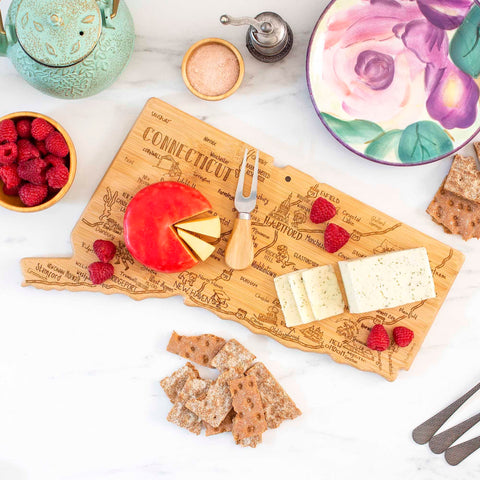 A Totally Bamboo Destination Connecticut Cutting Board, measuring 16-1/2" x 9-1/2" and featuring laser-engraved artwork, displays cheese, a cheese knife, raspberries, and crackers. Adjacent to it are a teapot, salt cellar, a floral plate, and cutlery on a white marble surface.