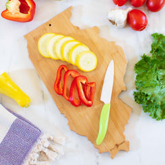 A Totally Bamboo Destination Illinois Cutting Board, measuring 16-1/2" x 9-1/4", serves as the backdrop for showcasing sliced yellow squash and red bell pepper. A knife with a green handle is resting beside it. Surrounding this Illinois-shaped cutting board are whole vegetables, such as a tomato, garlic, lettuce, and peppers.