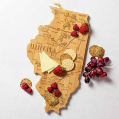 The Destination Illinois Cutting Board by Totally Bamboo, measuring 16-1/2" x 9-1/4", features a bamboo surface with laser-engraved artwork of Illinois city names. It is beautifully topped with strawberries, cheese slices, crackers, and grapes on a white surface.