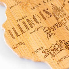 The Destination Illinois Cutting Board by Totally Bamboo, measuring 16-1/2" x 9-1/4", is expertly crafted into the shape of Illinois, highlighting laser-engraved artwork that includes the state outline, "ILLINOIS," a sketch of the capitol, floral motifs, and city names such as Quincy, Jerseyville, and Springfield.