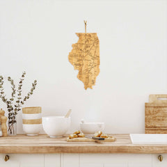 A cozy kitchen scene showcases a wooden Illinois map-themed cutting board from Totally Bamboo hanging on a white wall. Below, there's a countertop adorned with white bowls, decorative jars, and a vase filled with eucalyptus branches resting on the Destination Illinois Cutting Board. A few cookies are placed on a plate nearby.