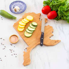 The Totally Bamboo "Destination Alaska Cutting Board," measuring 13-3/8" x 13-1/2", showcases the state of Alaska with a bamboo cutting surface. It's beautifully decorated with a knife, sliced zucchini, and yellow squash. Surrounding it on the marble surface are whole zucchini, tomatoes, lettuce, peppercorns, garlic, a blue-patterned dish, and a small bowl of spices.