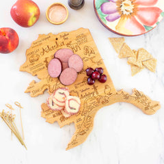 The Totally Bamboo Destination Alaska Cutting Board, measuring 13-3/8" x 13-1/2", elegantly displays sausage slices, cherries, and rolled appetizers on a marble surface. With apples, woven crackers, a colorful bowl, skewers, and a pepper shaker surrounding it, this state-shaped board beautifully enhances any setting.