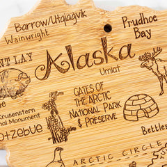 The Destination Alaska Cutting Board by Totally Bamboo is a wooden plaque shaped like the state of Alaska, adorned with laser-engraved designs of iconic landmarks and symbols such as a moose, igloo, and the Arctic Circle. It also features names like "Barrow" and "Gates of the Arctic National Park & Preserve," enhancing its distinctive appeal. The cutting board measures 13-3/8" x 13-1/2".