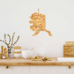 A kitchen scene features a light wooden countertop with white bowls, a bamboo cutting surface from the Totally Bamboo Destination Alaska Cutting Board collection, and salt and pepper shakers. Above the countertop is a wooden wall decoration shaped like the state of Alaska with laser-engraved artwork and text.