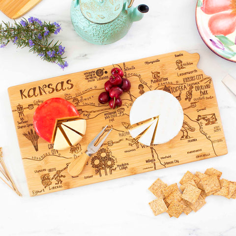 The Totally Bamboo Destination Kansas Cutting Board, measuring 16-3/4" x 9-1/4", features laser-engraved city names and landmarks. Decorated with two varieties of cheese, a cheese fork, crackers, grapes, and a ceramic serving pot, it sits gracefully on a marble surface next to a plant and plate.