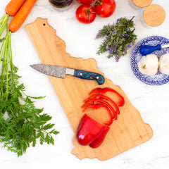 A knife and sliced red bell pepper sit on the Destination New Hampshire Cutting Board by Totally Bamboo, which showcases intricate laser-engraved artwork. Fresh carrots, tomatoes, garlic, herbs, and spices are arranged around the board on a marble surface.