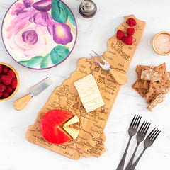 A Totally Bamboo Destination New Hampshire Cutting Board, measuring 9" x 17", features laser-engraved artwork and is adorned with assorted cheeses and raspberries. Accompanying it are crackers, cutlery, a painted floral plate, and a small bowl of salt on a white marble surface.