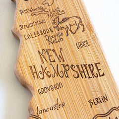 The Totally Bamboo Destination New Hampshire Cutting Board, measuring 9" x 17", is precisely crafted with a bamboo cutting surface and laser-engraved to depict the outline of New Hampshire. It features place names like Pittsburg, Stewartstown, and Lancaster alongside an illustration of a moose, with "New Hampshire" stylishly carved in the center.