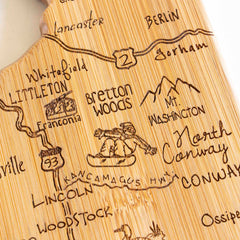 The Totally Bamboo Destination New Hampshire Cutting Board, measuring 9" x 17", is uniquely crafted from bamboo in the shape of the state and features a laser-engraved map highlighting locations such as North Conway, Berlin, and Bretton Woods. It is adorned with an illustration of a snowboarder and showcases mountain peaks like "Mt. Washington" and "Kancamagus Hwy," along with detailed representations of Routes 2 and 93.