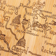 The Totally Bamboo Destination New Hampshire Cutting Board, measuring 9" x 17", features intricate laser-engraved illustrations of towns and landmarks across New Hampshire, USA, highlighting places like Concord and Lake Winnisquam. The board is further adorned with images of a motorcycle, a tree, and a domed building.