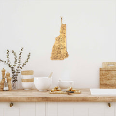 A cozy kitchen scene features a wooden countertop with the Destination New Hampshire Cutting Board by Totally Bamboo as the bamboo cutting surface, alongside a white mixing bowl and delicious pastries. Charming decorative jars and eucalyptus branches enhance the ambiance, while a laser-engraved wooden map of New Hampshire is proudly displayed on the wall above.