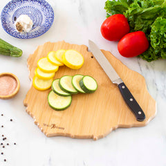 A Destination Kauai Cutting Board by Totally Bamboo is placed on a marble countertop, adorned with sliced zucchini, a black-handled knife, tomatoes, lettuce, a blue plate containing garlic and zucchini, and a bowl of salt sprinkled with black peppercorns.