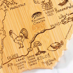 The Totally Bamboo Destination Kauai Cutting Board, measuring 9-3/4" x 12-1/2", features laser-engraved artwork depicting a map of Kauai, Hawaii, adorned with illustrations such as a rooster and a fish, as well as place names including Lihue and Poipu Beach Park.