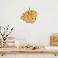 A cozy kitchen showcases a wooden map of Kauai from the Totally Bamboo Destination Series on a white wall. Beneath it, a light wood countertop with the Totally Bamboo Destination Kauai Cutting Board, measuring 9-3/4" x 12-1/2", is adorned with white bowls, a mortar and pestle, wooden utensils, and a small plant in a vase. This arrangement creates an inviting and warm ambiance.