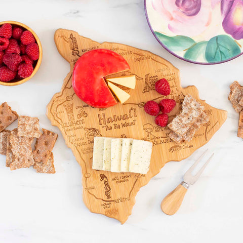 The Totally Bamboo Destination Hawaii Cutting Board, measuring 13-1/4" x 11-1/2", beautifully displays its laser-engraved artwork as it holds Brie, Gouda, raspberries, and crackers. Additional crackers and raspberries complement the scene on a decorative plate nearby, accompanied by an open cheese knife.