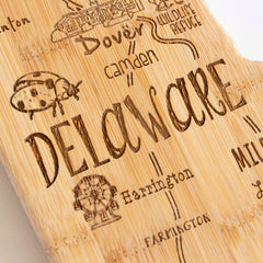 The Destination Delaware Cutting Board by Totally Bamboo is a laser-engraved, state-shaped board measuring 16-1/2" x 9-1/2", crafted from bamboo and featuring the word "Delaware," with delightful illustrations of locations such as Dover, Camden, and Harrington along with landmarks like a ladybug and a tree.