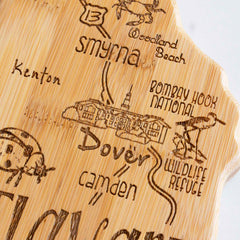 A detailed view of the Destination Delaware Cutting Board by Totally Bamboo, measuring 16-1/2" x 9-1/2", shows Delaware's outline with laser engravings of locations such as Dover, Smyrna, and Camden. It includes depictions of landmarks like Bombay Hook National Wildlife Refuge, adorned with natural motifs and a sketch of a building.