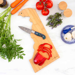 A wooden, state-shaped board intricately laser-engraved with artwork of Delaware holds a knife and sliced red bell pepper. Around it, whole red peppers, tomatoes, carrots with greens, a plate of garlic, and fresh herbs rest elegantly on the marble surface. The board is identified as the Destination Delaware Cutting Board by Totally Bamboo and measures 16-1/2" x 9-1/2".