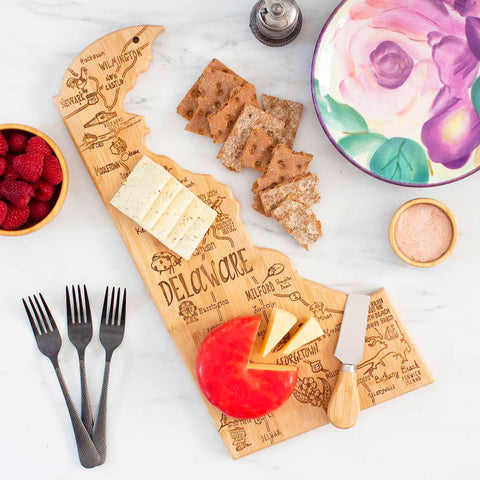 A Totally Bamboo Destination Delaware Cutting Board, measuring 16-1/2" x 9-1/2", showcases laser-engraved artwork and holds an array of crackers, cheese, and a wheel of cheese elegantly displayed on a marble surface. Accompanying the setup are raspberries in a bowl, a floral-patterned plate, a dish of salt with its shaker, along with a knife and four forks.