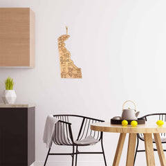 A minimalist kitchen features a light wood table adorned with a teapot, cups, and lemons. Black wireframe chairs complement the white wall where a laser-engraved map of Delaware artfully hangs. A Totally Bamboo Destination Delaware Cutting Board, measuring 16-1/2" x 9-1/2", rests beside a potted plant on the counter, enhancing the serene setting.