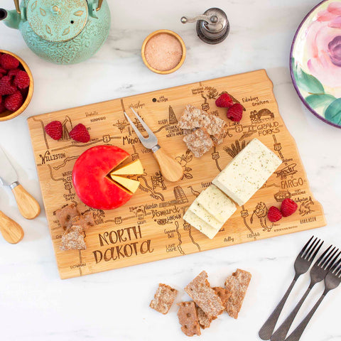 The Destination North Dakota Cutting Board by Totally Bamboo, measuring 9-3/4" x 16", is crafted from bamboo and features an etched map of North Dakota. It gracefully displays cheese and crackers with accompanying cheese knives, alongside a teapot, a bowl of raspberries, a salt bowl, a floral plate, and four forks on the marble surface.