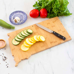 A Totally Bamboo Destination South Dakota Cutting Board, measuring 9-3/4" x 16", is artfully arranged with sliced green and yellow squash. Nearby, a knife rests beside fresh tomatoes, lettuce, zucchini, salt, pepper, and a plate with garlic on a white marble countertop.