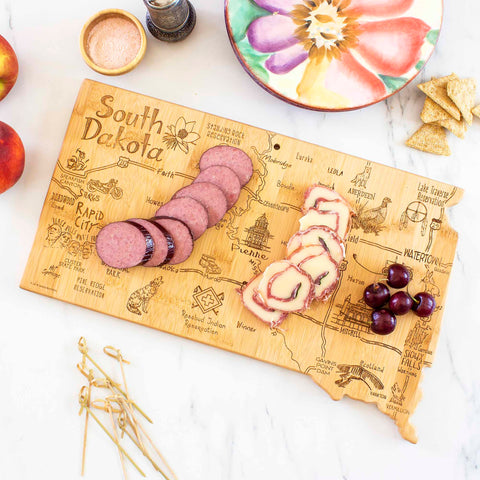 The Destination South Dakota Cutting Board by Totally Bamboo, measuring 9-3/4" x 16", serves as a charming wooden charcuterie board shaped like South Dakota, displaying sliced sausage, cheese, and cherries. It is laser-engraved with cities and landmarks and elegantly accompanied by a colorful plate, crackers, and seasoning.