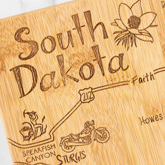 Introducing the Totally Bamboo Destination South Dakota Cutting Board, measuring 9-3/4" x 16". This state-shaped board features a detailed map of South Dakota with engravings including decorative elements like a flower and charming illustrations of a motorcyclist and gold panner. Key towns such as Spearfish Canyon and Sturgis are marked along Highway 212.