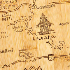 The Totally Bamboo Destination South Dakota Cutting Board, measuring 9-3/4" x 16", features a wooden map carving highlighting parts of the state, including Pierre, Gettysburg, and Mission Ridge. It showcases an illustration of the Pierre State Capitol building alongside an old-fashioned car and I-90 highway on this state-shaped board.