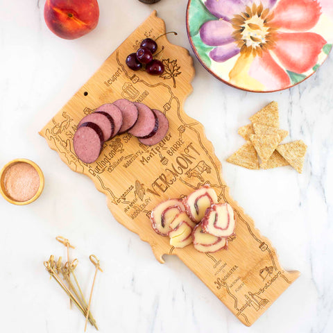 A Totally Bamboo Destination Vermont Cutting Board, measuring 16" x 10" and featuring a laser-engraved design, holds slices of salami and cheese. Nearby on the marble surface are cherries, a peach, a bowl of pink salt, a floral plate, crackers, and skewers.