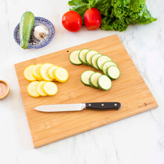 A Totally Bamboo Destination Wyoming Cutting Board, measuring 11" x 14-1/4", features sliced yellow and green zucchini, a knife, and a small bowl. In the background on a marble surface are fresh lettuce, tomatoes, zucchini, and garlic, enhanced by laser-engraved artwork.