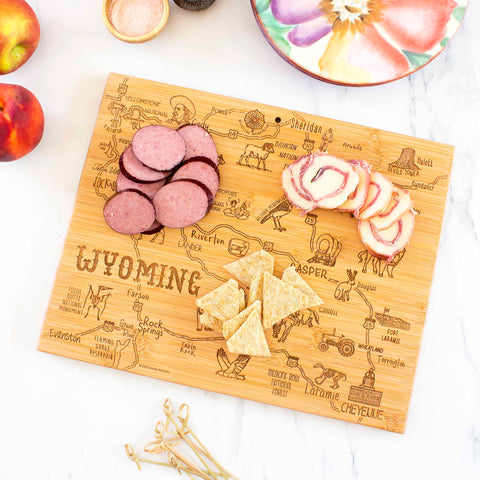 Slices of salami, cheese, and crackers rest on the Totally Bamboo Destination Wyoming Cutting Board, measuring 11" x 14-1/4", which is shaped like the state and engraved with a map. Nearby, two whole apples and a vibrant ceramic plate adorn the marble countertop.