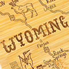 The Totally Bamboo "Destination Wyoming Cutting Board," measuring 11" x 14-1/4", showcases a laser-engraved, hand-drawn map of Wyoming on a state-shaped board. The design features illustrated animals like a deer and an eagle, as well as marked highways connecting cities such as Lander, Rock Springs, and Green River. The word "WYOMING" is prominently displayed.