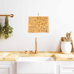 In a cozy kitchen, the Totally Bamboo Destination Wyoming Cutting Board, measuring 11" x 14-1/4", hangs artfully on a white wall above a copper sink. Fresh herbs dangle nearby, adding greenery, while the bamboo cutting board serves both as an attractive décor piece and a functional surface next to the wooden utensil holder on the countertop.