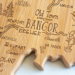 The Totally Bamboo Destination Maine Cutting Board, measuring 15" x 10-1/2", is designed in the shape of Maine and features laser-engraved artwork that highlights place names like Bangor, Newport, Bar Harbor, and Belfast. Decorative wavy lines and trees add unique touches to this piece.