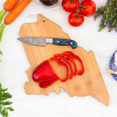 A rooftop view features the Destination Maine Cutting Board by Totally Bamboo, measuring 15" x 10-1/2", artfully laden with sliced red bell peppers next to a patterned knife. Fresh carrots, tomatoes, and herbs surround the board, with glimpses of a plate and bottle nearby.