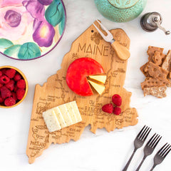 The Totally Bamboo Destination Maine Cutting Board, measuring 15" x 10-1/2", features a charming shape of the state and holds cheese slices, a wheel of cheese, and raspberries. It is beautifully adorned with laser-engraved artwork and surrounded by crackers, a floral plate, a turquoise bowl, and four forks on a white surface.