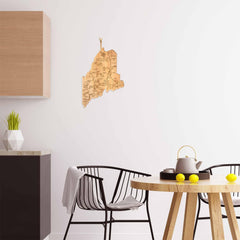 In this modern dining area, a round wooden table is surrounded by black chairs. A teapot with cups and lemons add charm, while the Destination Maine Cutting Board from Totally Bamboo complements the geometric plant pot on the marble countertop. The wooden map of Maine adorns the white wall.