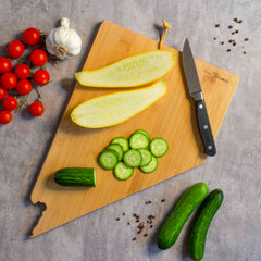 The Destination Nevada Cutting Board by Totally Bamboo, measuring 10" x 15-1/2", showcases sliced and halved cucumbers, a knife, cherry tomatoes, a garlic bulb, and scattered peppercorns against a gray background. The laser-engraved artwork enhances its charm, making it an ideal addition to any kitchen setting.