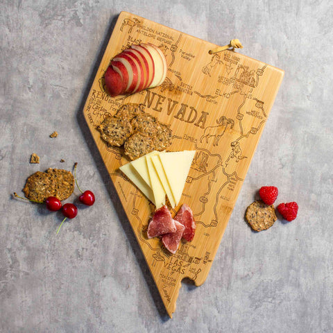 A Destination Nevada Cutting Board by Totally Bamboo, measuring 10" x 15-1/2", features a laser-engraved artwork and is artfully arranged with sliced red-skinned apples, crackers, cheese, and cured meat. Beside it are cherries, raspberries, and additional crackers on a gray surface.