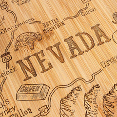 Crafted from wood, the Totally Bamboo Destination Nevada Cutting Board, measuring 10" x 15-1/2", features a state-shaped design with laser-engraved details. It showcases highways, towns like Winnemucca and Carlin, a tortoise icon near Battle Mountain, and mountain ranges. The word "Nevada" is prominently carved at the center of this unique artwork.