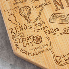 The Totally Bamboo Destination Nevada Cutting Board, measuring 10" x 15-1/2", is beautifully designed with a laser-engraved map showcasing Nevada's cities and landmarks such as Reno, Carson City, and Lake Tahoe. This bamboo cutting surface also features unique artwork including a hot air balloon and a star.