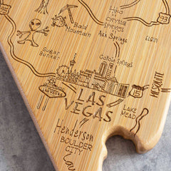 Introducing the Totally Bamboo Destination Nevada Cutting Board, a 10" x 15-1/2" bamboo cutting surface uniquely shaped like the state of Nevada. It features exquisite laser-engraved artwork of Las Vegas, Mesquite, Bald Mountain, and Lake Mead. Additional decorative details include a UFO depiction, a cactus illustration, and an intricately etched city skyline.