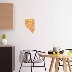 A modern kitchen showcases a laser-engraved wall decoration in the shape of Nevada using the Destination Nevada Cutting Board from Totally Bamboo. The scene is completed with a black chair draped with a gray blanket, alongside a round wooden table adorned with a teapot and three yellow apples. A light wooden cabinet and a white potted plant enhance the décor.