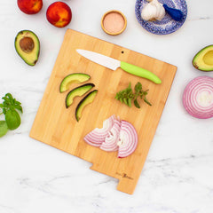 Blank back side of New Mexico state shaped bamboo board, holding sliced vegetables and paring knife, on white marble countertop with vegetables surrounding